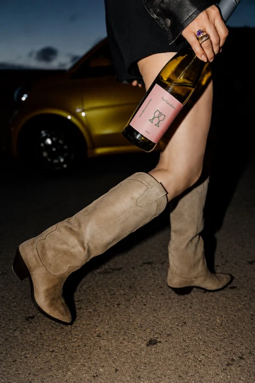 Someone in suede cowboy boots carrying a bottle of rosé at night